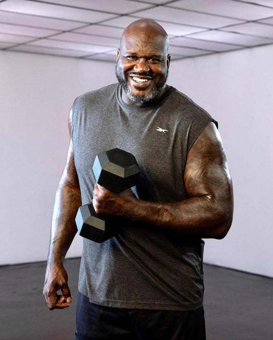 Shaquille O'Neal smiling while holding a black dumbbell in a gym