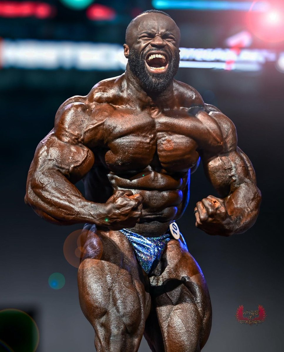 Samson Dauda posing under Olympia stage lights with massive muscularity and conditioning