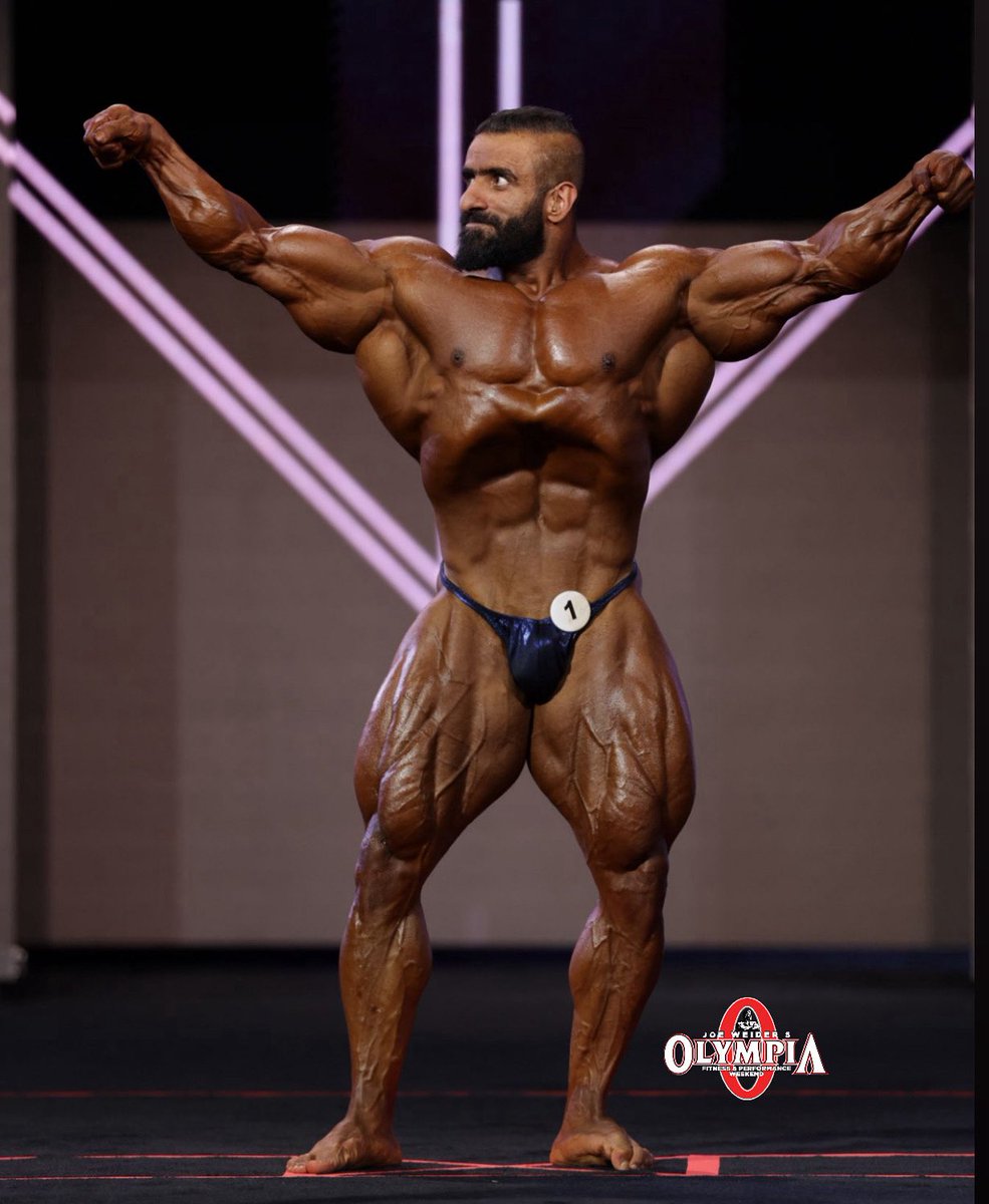 Hadi Choopan standing on a bodybuilding stage with his arms raised and visibly defined chest, abs and quadriceps