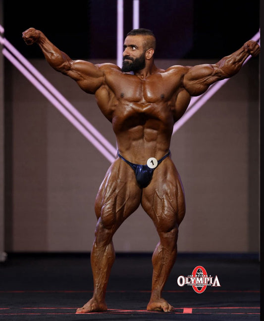 Hadi Choopan standing on a bodybuilding stage with his arms raised and visibly defined chest, abs and quadriceps