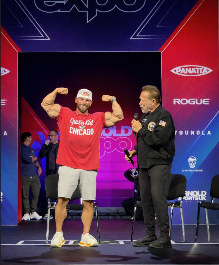 Joey Swoll posing in a gym with red weight machines, wearing a red cap and white shorts.