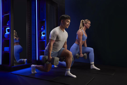 Official Solidcore Ascend Strong50 participants performing heavy dumbbell lunges in a dark blue-lit fitness studio; editorial news photo, real people, no graphic overlays.