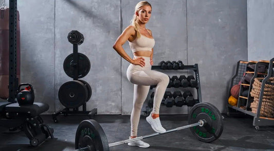 Heidi Somers posing in a beige gym outfit with one foot on a barbell beside dumbbell racks