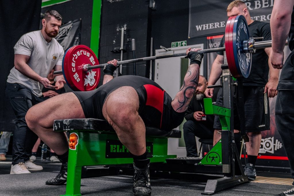 Powerlifter on a flat competition bench pressing a heavy barbell with red plates as spotters stand nearby