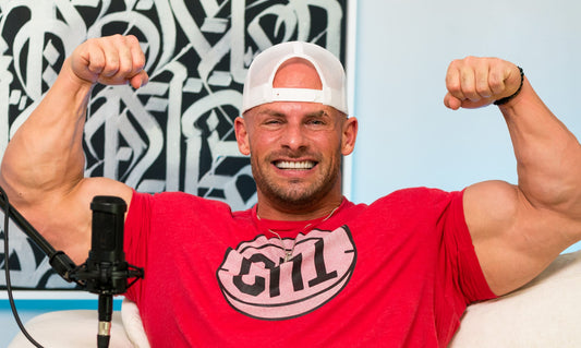 Joey Swoll smiling and flexing in a red shirt during a studio interview