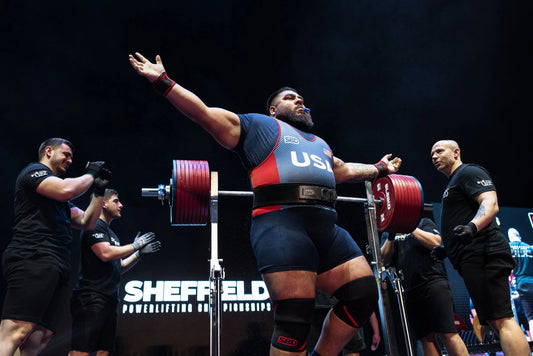 Jesus Olivares celebrating on a powerlifting platform beside a heavily loaded squat rack