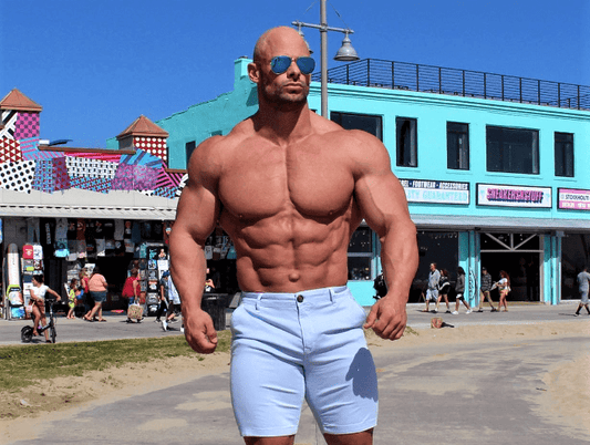 Joey Swoll wearing a black Gym Positivity Nation tank top and flexing both arms in a gym