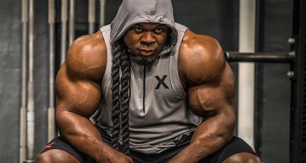 Kai Greene seated in a gym wearing a sleeveless hoodie, showing his powerful arms and focused expression