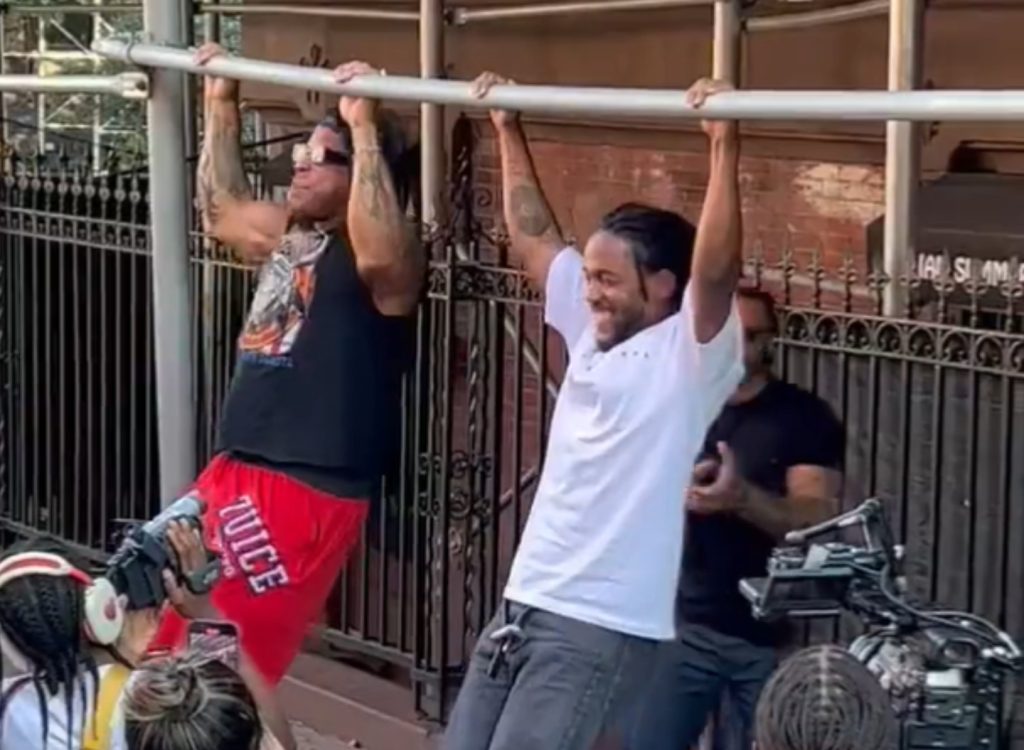 Kendrick Lamar hanging from a street pull-up bar alongside another participant in Harlem