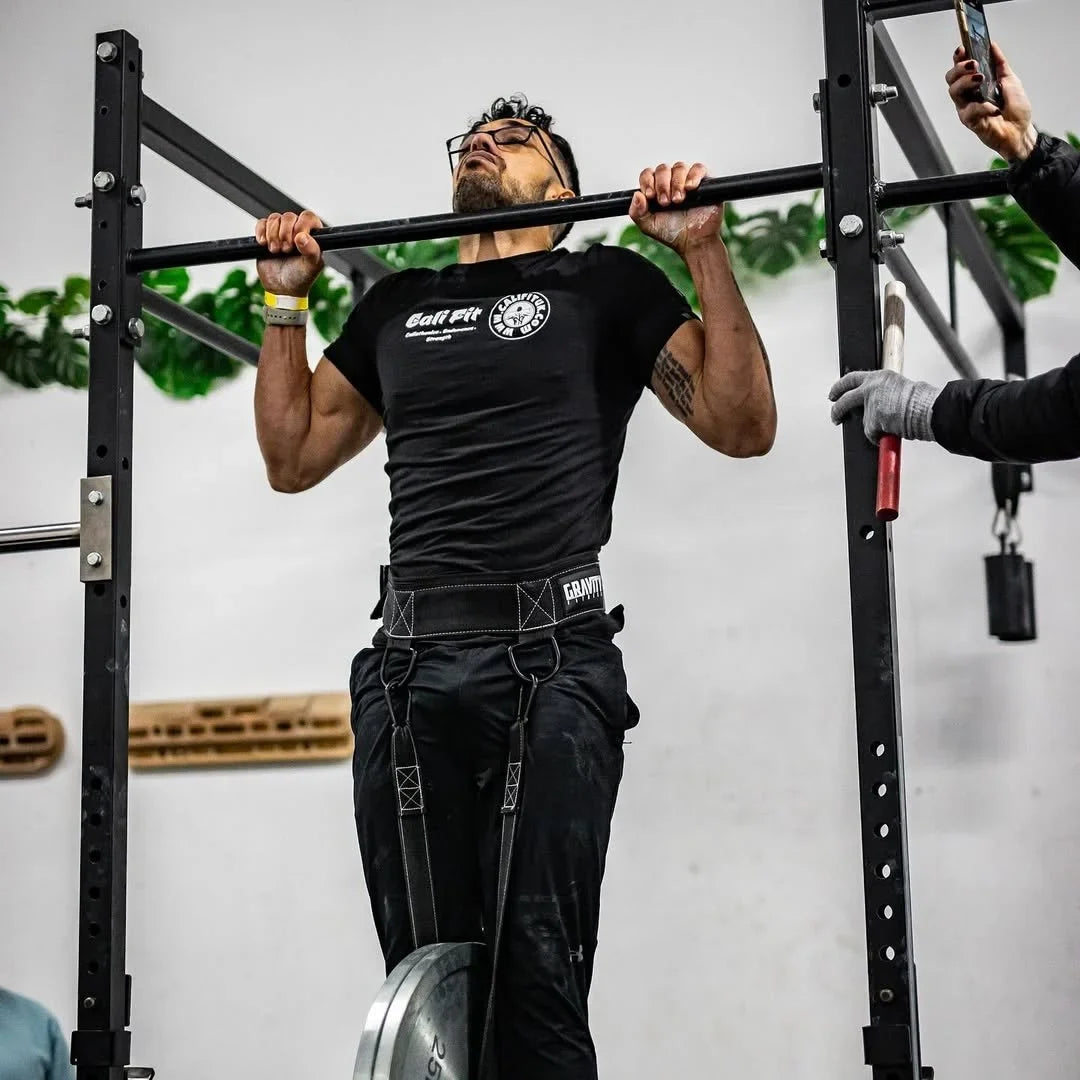 Male athlete performing a heavy weighted pull-up with a weight plate hanging from a dip belt in a gym.
