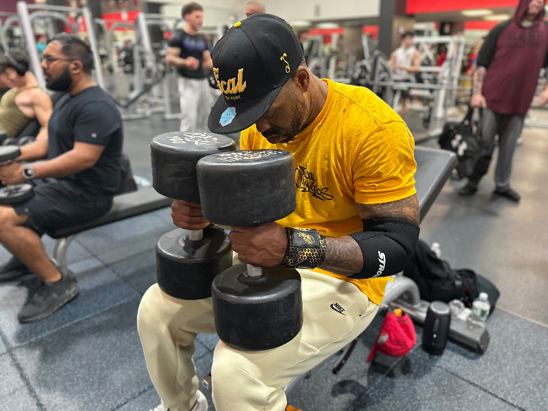 Method Man seated in a gym holding heavy dumbbells while wearing a yellow Tical Athletics shirt