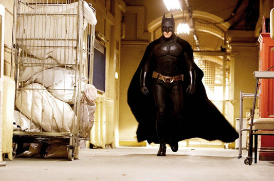 Christian Bale as Batman walking in the black armored Batman Begins suit with a broad chest and sweeping cape