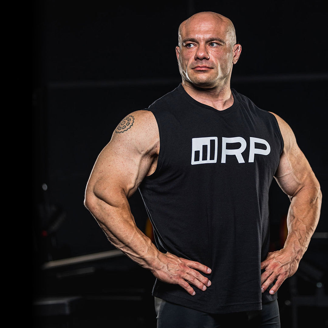 Mike Israetel posing in a black RP Strength tank top inside a dim gym