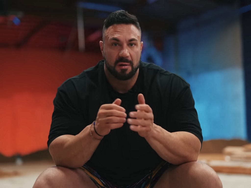 Noel Deyzel seated in a dark gym, looking toward the camera and gesturing as he speaks