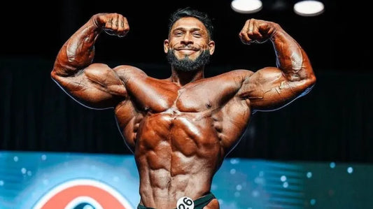 Ramon Dino performing a front double-biceps pose on a bodybuilding stage, showing his muscular chest, arms and midsection