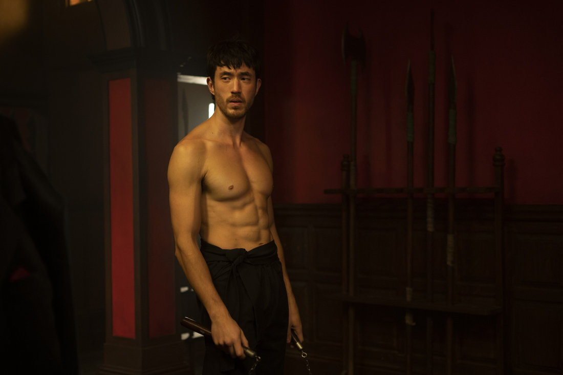 Andrew Koji as Ah Sahm shirtless and holding nunchaku in a dark martial-arts training room