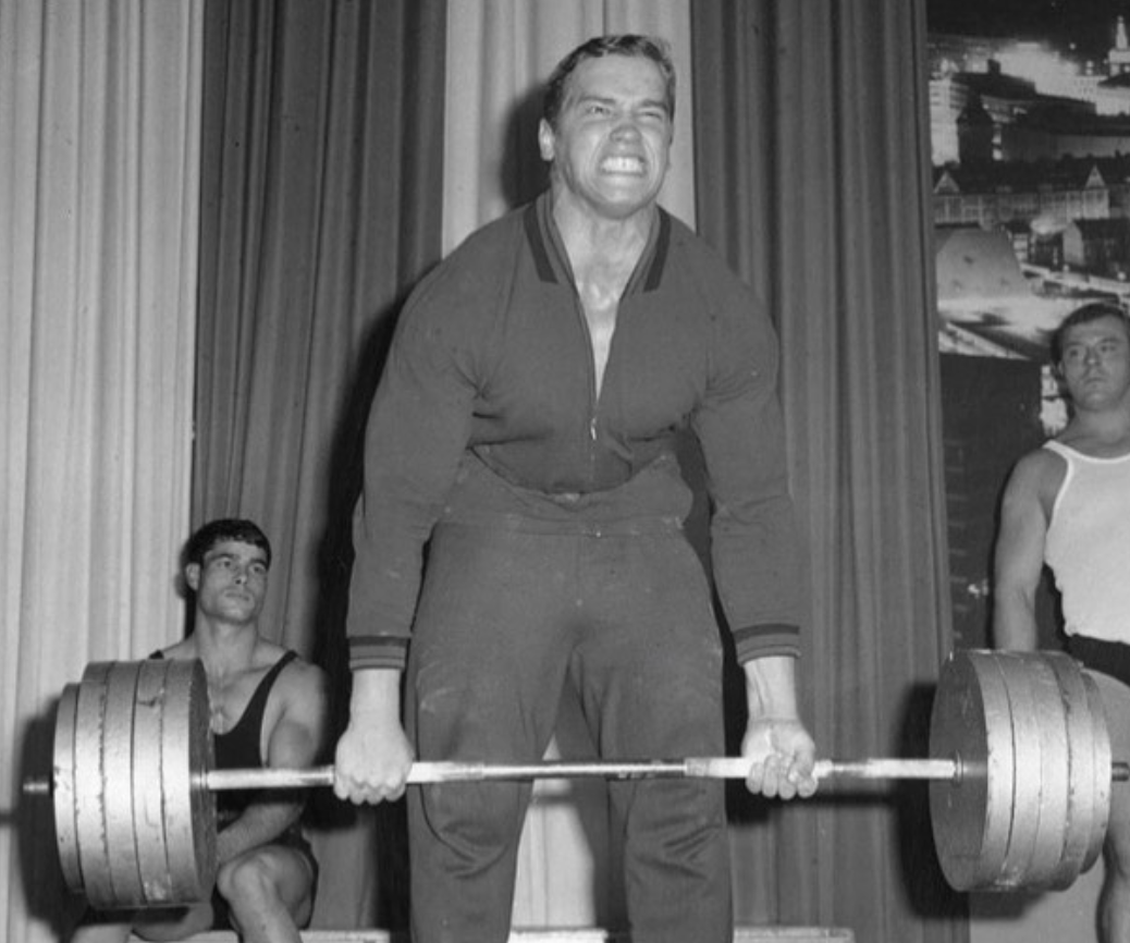 Young Arnold Schwarzenegger completing a heavy barbell deadlift on a vintage competition stage