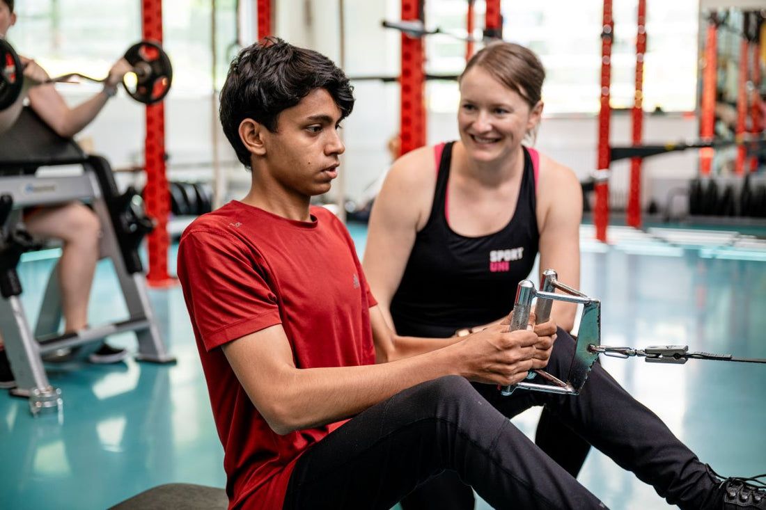 Beginner practicing a seated cable row with a trainer coaching nearby in a university gym