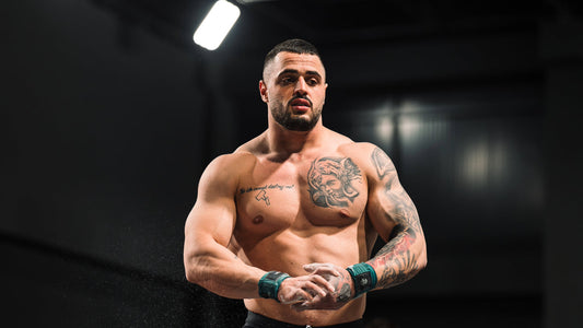 Karlos Nasar preparing for a lift, shirtless with tattooed shoulders and chalked hands under dark training lights