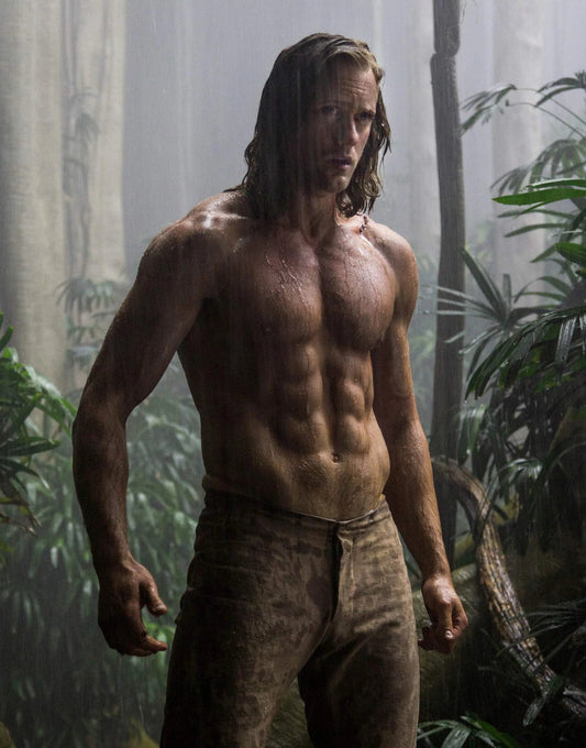 Alexander Skarsgård as Tarzan standing shirtless in a green jungle with broad shoulders and defined torso
