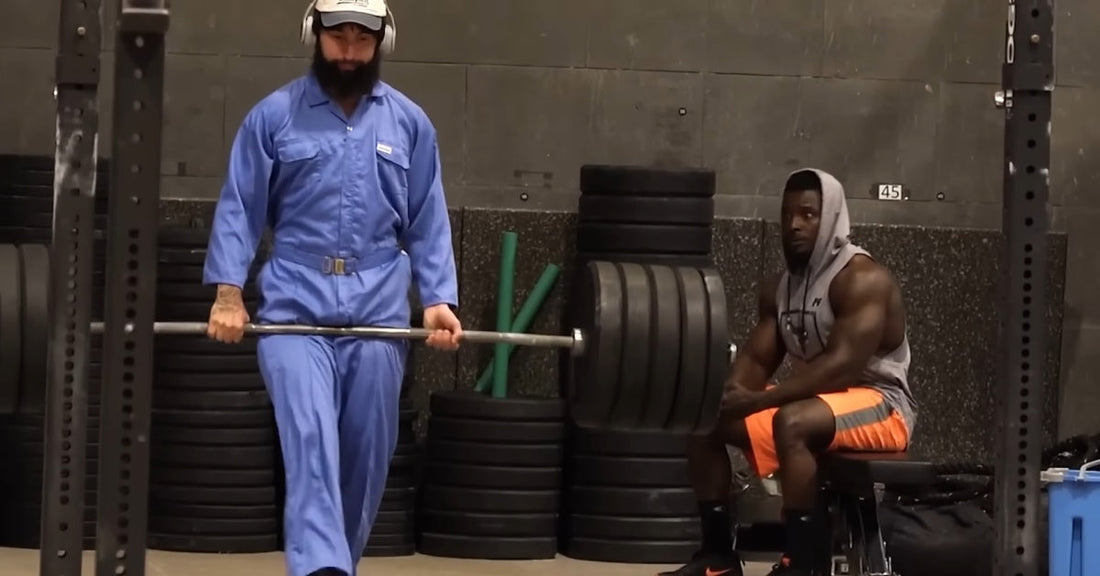 Anatoly in blue cleaner overalls deadlifting a loaded barbell beside two larger gym-goers