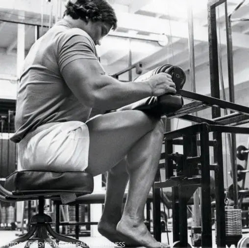 Arnold Schwarzenegger performing a seated calf raise in a vintage bodybuilding gym