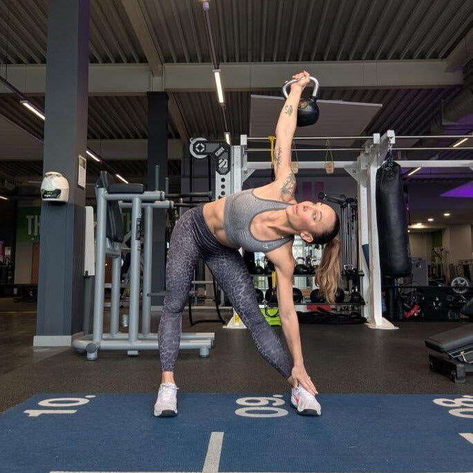 Woman demonstrating a controlled kettlebell windmill with one arm locked overhead and her opposite hand reaching toward the floor
