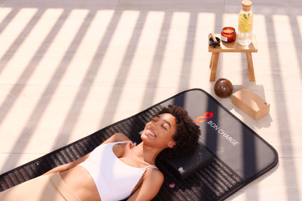 Woman reclining on a black BON CHARGE infrared PEMF mat in a sunlit room