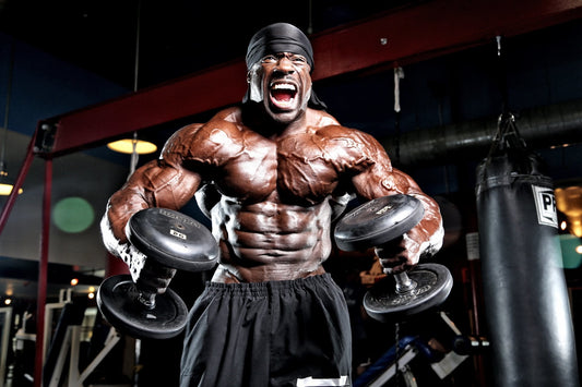 Kali Muscle flexing with massive pectorals and arms while holding dumbbells in a gym