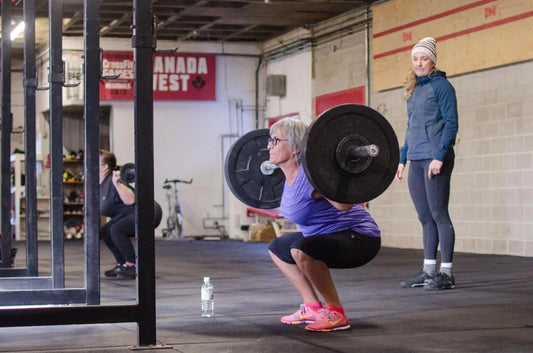 An older woman performs a supervised barbell squat in a gym