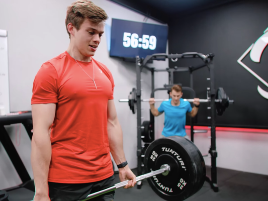 Stan Browney in a red training shirt holding a loaded barbell in a gym during a timed strength challenge