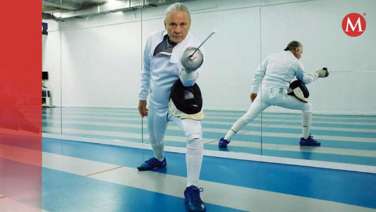Bruce Dickinson in white fencing clothes lunging with his blade on a blue-and-white indoor fencing piste