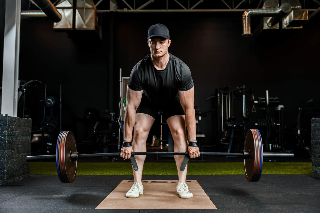 Male athlete preparing a loaded barbell deadlift on a gym platform, wearing a black shirt and cap
