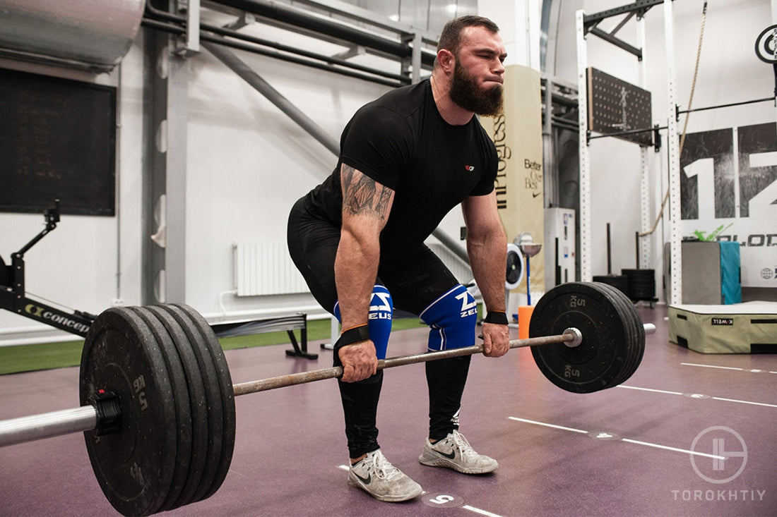Strong athlete in a black shirt and knee sleeves pulling a heavily loaded barbell from a gym platform