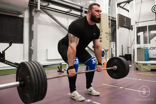 Strong athlete in a black shirt and knee sleeves pulling a heavily loaded barbell from a gym platform