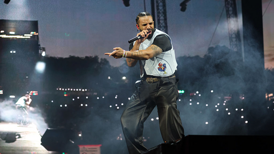 Drake performing at Manchester Co-op Live, pointing toward the audience with a microphone, in a white vest and dark pants.