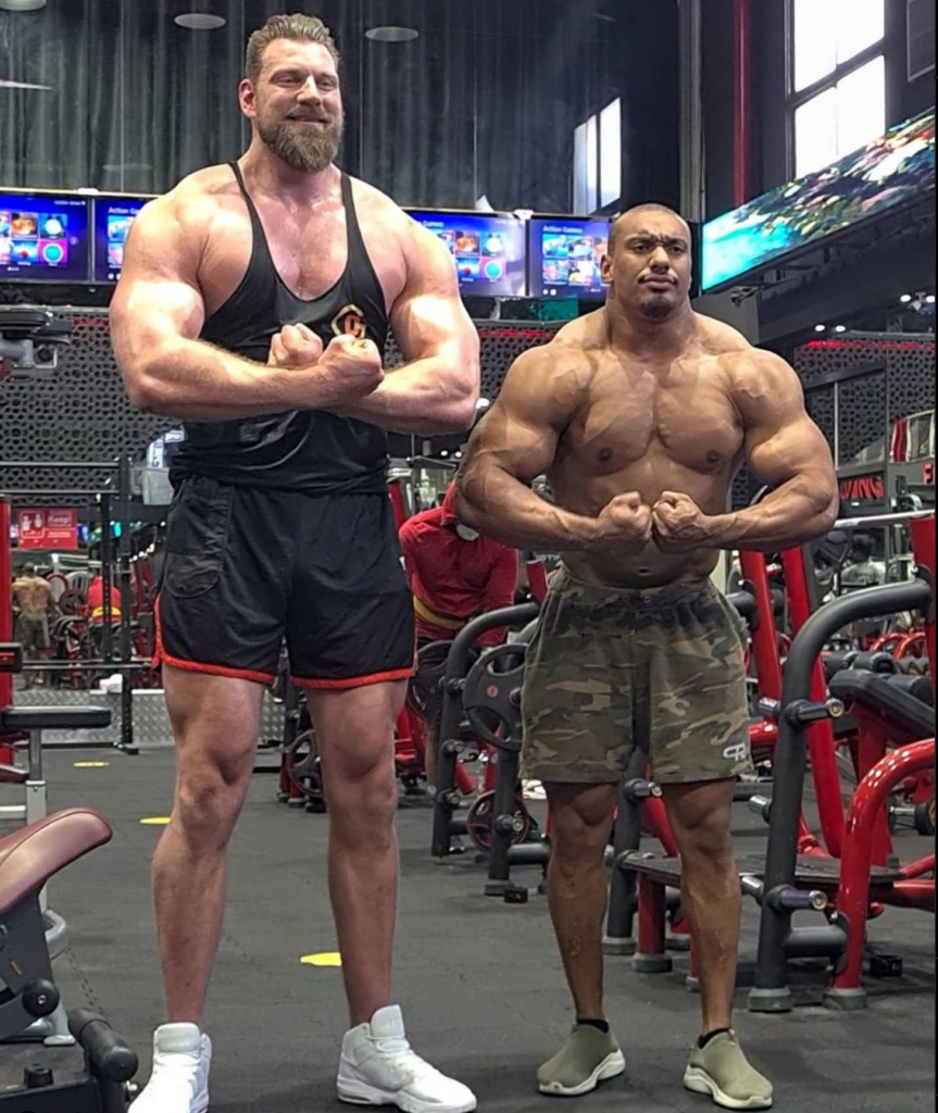 Olivier Richters, the Dutch Giant, flexes beside another muscular bodybuilder in a gym, showing the striking difference in height and frame size.