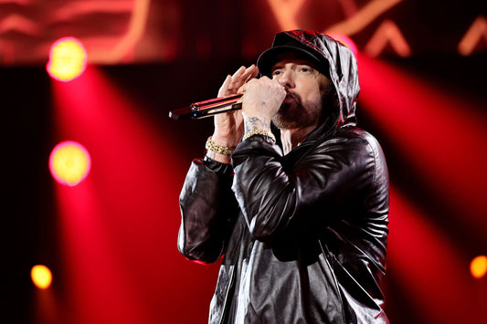 Eminem performing into a microphone in a black hooded jacket under red concert lights