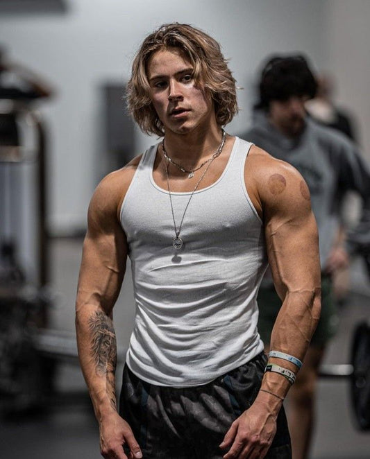 Alex Eubank in a white tank top standing in a gym, showing his athletic upper-body physique