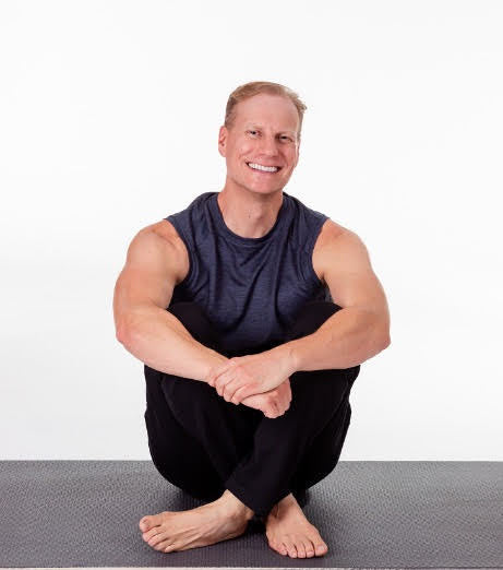 Sean Vigue, a muscular male Pilates instructor, seated on an exercise mat in a sleeveless top