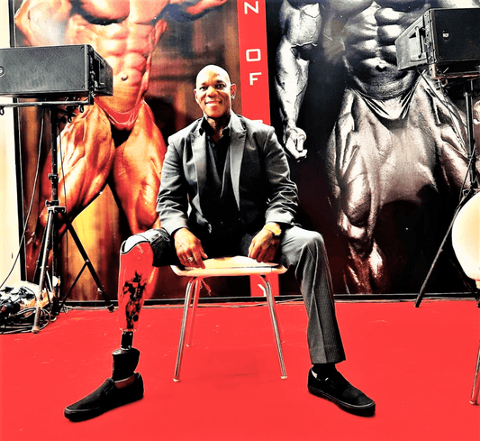 Flex Wheeler seated at a bodybuilding event in a dark suit, with his right prosthetic leg visible
