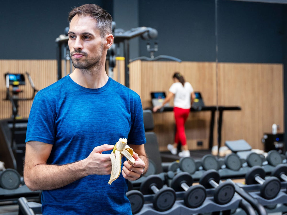 An athlete in a blue gym shirt holding a peeled banana beside dumbbell racks