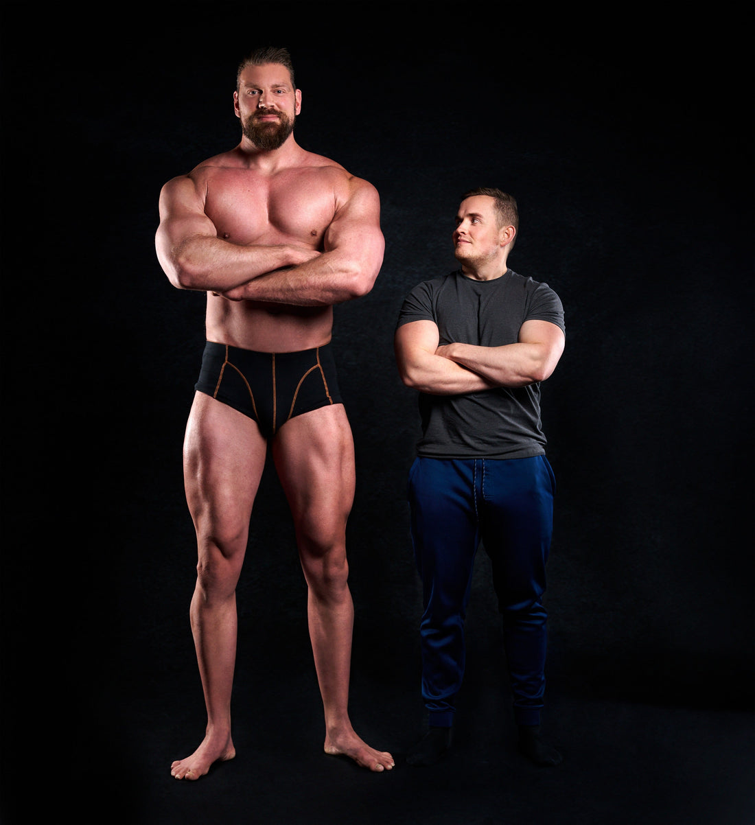 Olivier Richters standing shirtless with folded arms beside a shorter muscular man against a black studio backdrop