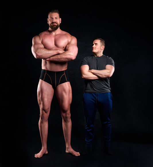 Olivier Richters standing shirtless with folded arms beside a shorter muscular man against a black studio backdrop