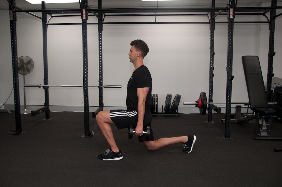 Male personal trainer performing a dumbbell split squat in a gym with front knee bent and rear knee near the floor.