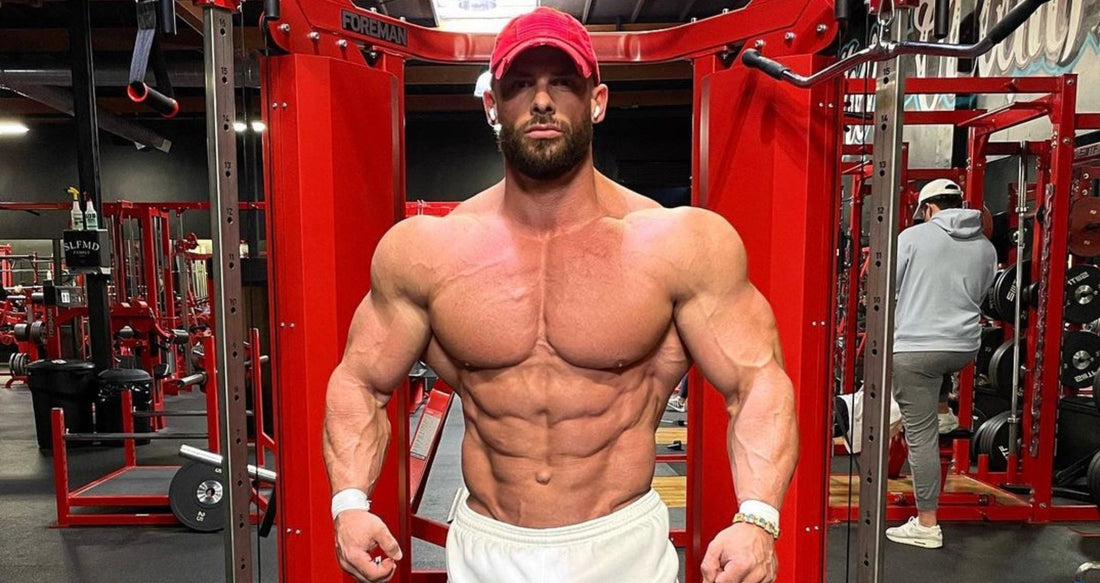 Joey Swoll posing shirtless in a red cap among red gym strength machines