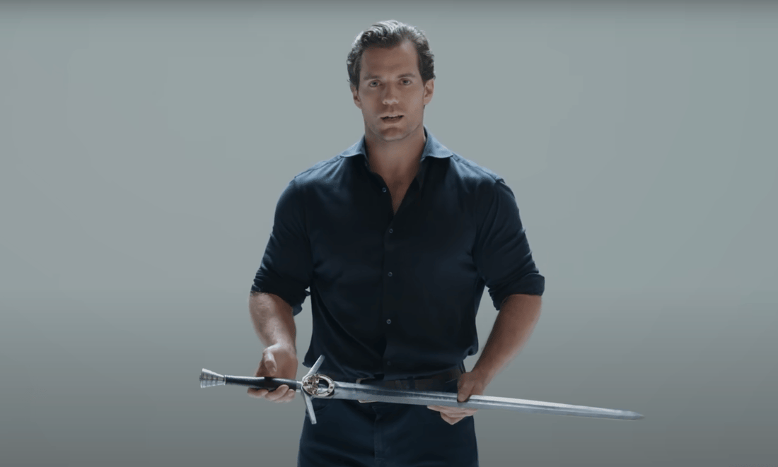 Henry Cavill holding a longsword in a dark shirt against a plain backdrop