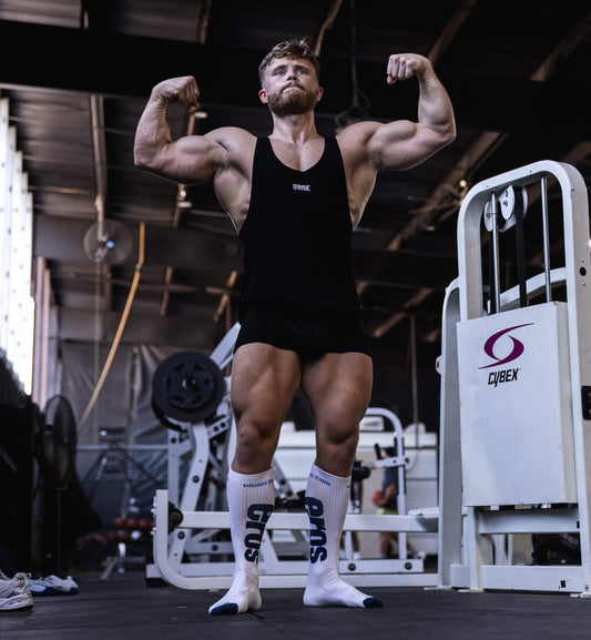 Jeff Nippard flexing his arms and legs in a black tank top in a weight-training gym