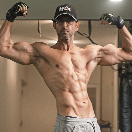 Hrithik Roshan flexing muscular arms and defined abs in a gym wearing a black cap