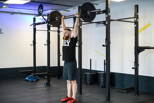 Bearded athlete standing inside a gym rack with a loaded barbell locked out overhead